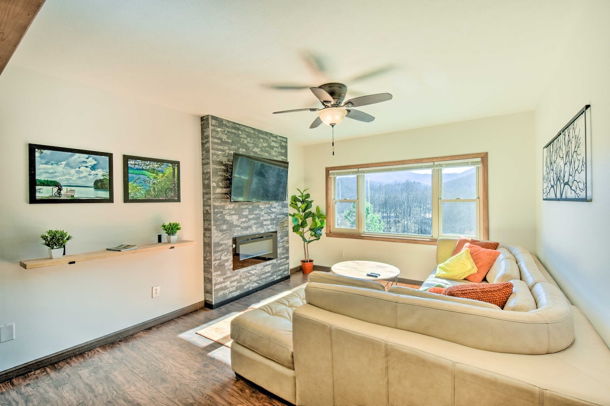 A spacious living area features a large, light-colored sectional sofa in front of a wall-mounted television. Natural light floods in through the large windows, showcasing mountain views. A modern electric fireplace is positioned beneath the TV, complemented by potted plants and framed artwork on the walls.