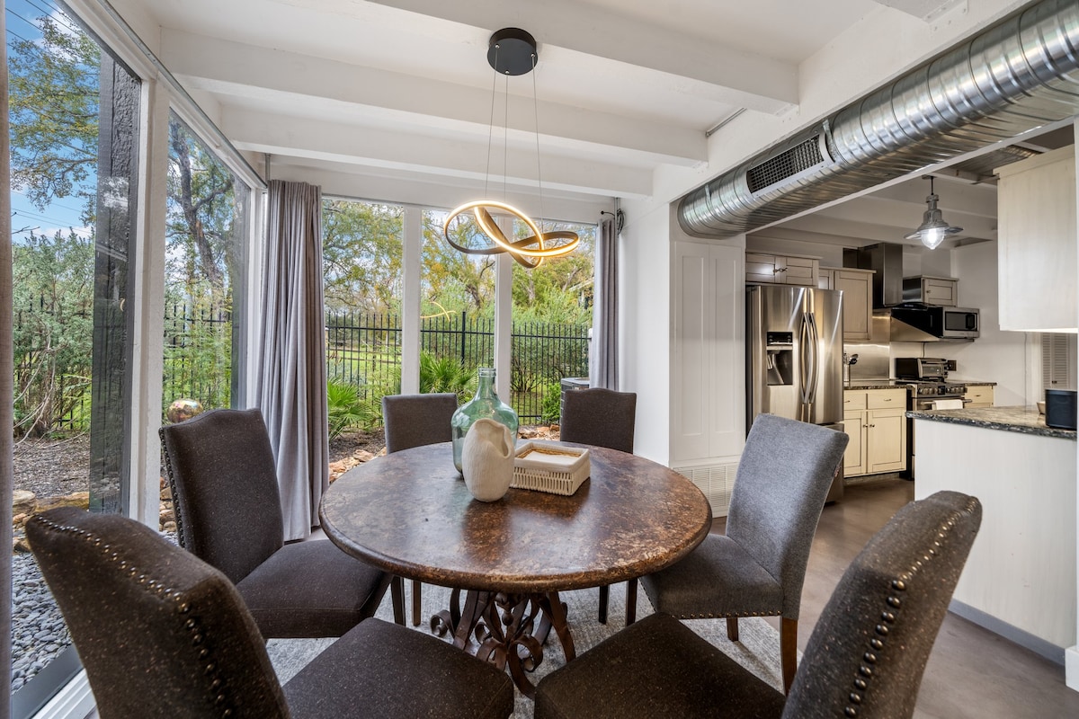 A round dining table with a rustic metal top is surrounded by six upholstered chairs. Large windows offer views of the outdoor landscape, while natural light fills the space. A modern light fixture hangs above the table, complementing the open design.