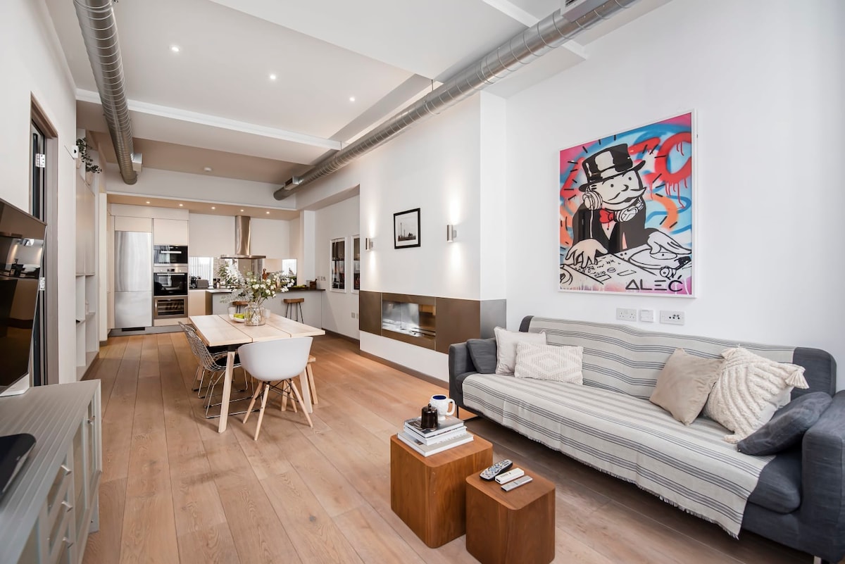 A bright living area features a modern dining table surrounded by white chairs and a stylish couch. A wall-mounted television is positioned across from the table. Artwork adorns one wall, with the kitchen visible in the background, showcasing sleek appliances and cabinetry.
