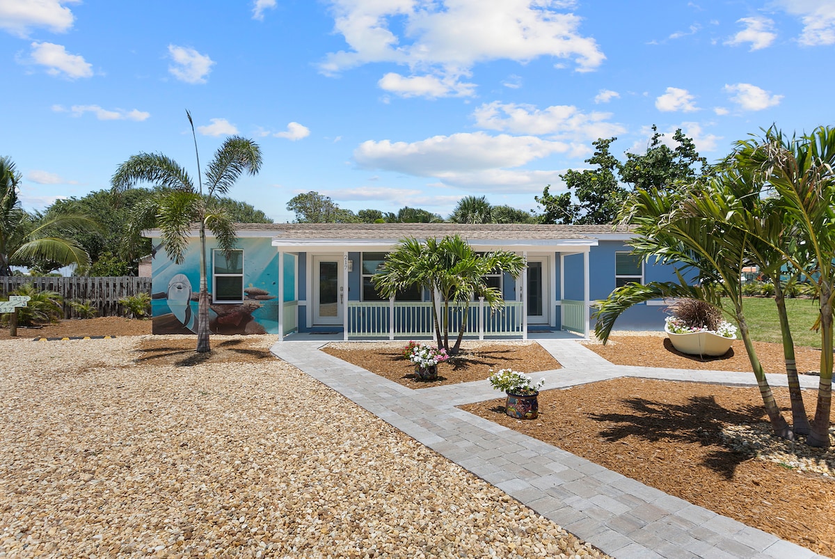 A charming duplex is set against a bright blue sky, showcasing a landscaped front yard with gravel and palm trees. The walkway is lined with pavers, leading to a welcoming porch with a white railing. Flower beds add color to the exterior.