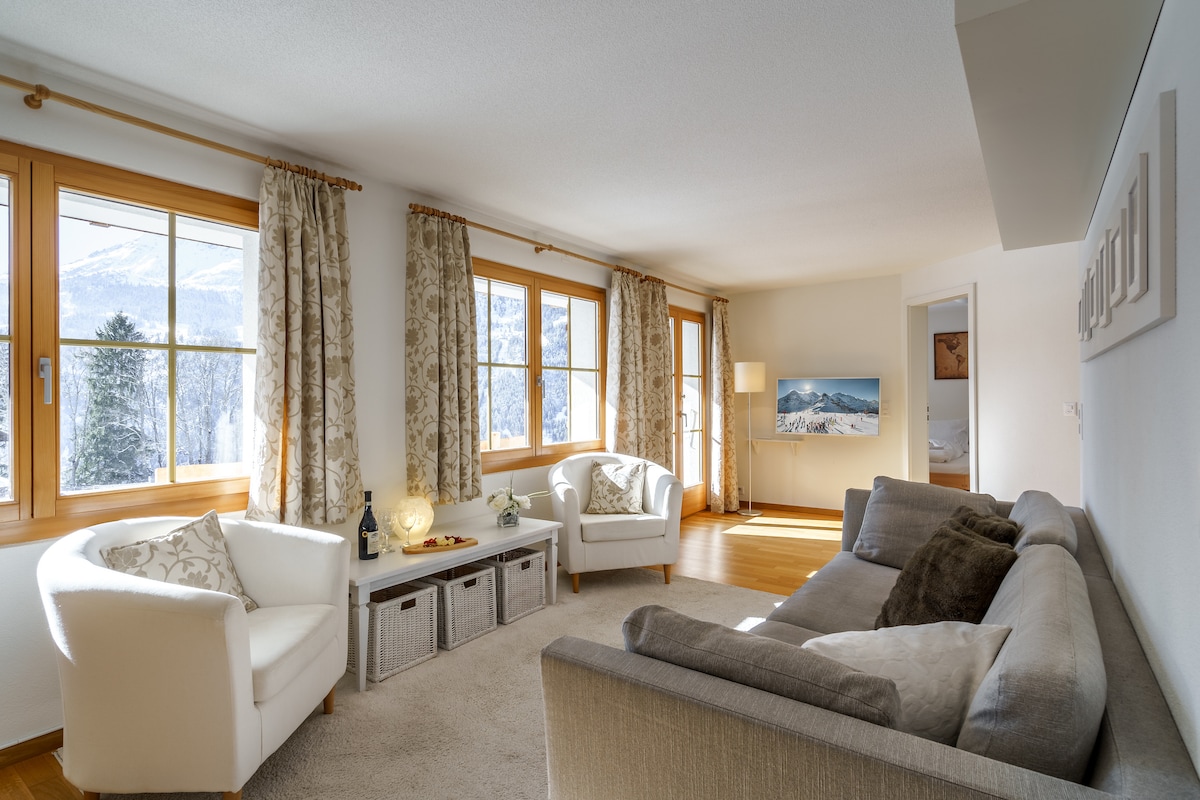 A bright living area features large windows framing mountain views and natural lighting. Soft beige walls complement contemporary furnishings, including a spacious gray sofa and two white armchairs. Decorative curtains add subtle texture, while a low coffee table sits invitingly in the center of the room.