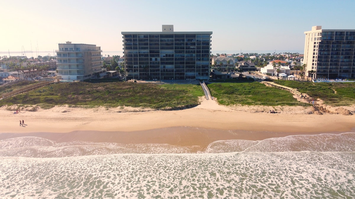 An aerial view captures the beachfront property along sandy shores, with the ocean's waves gently lapping at the beach. Nearby, the landscape reveals green lawns and pathways leading to the water, framed by low-rise buildings that offer direct shoreline access.