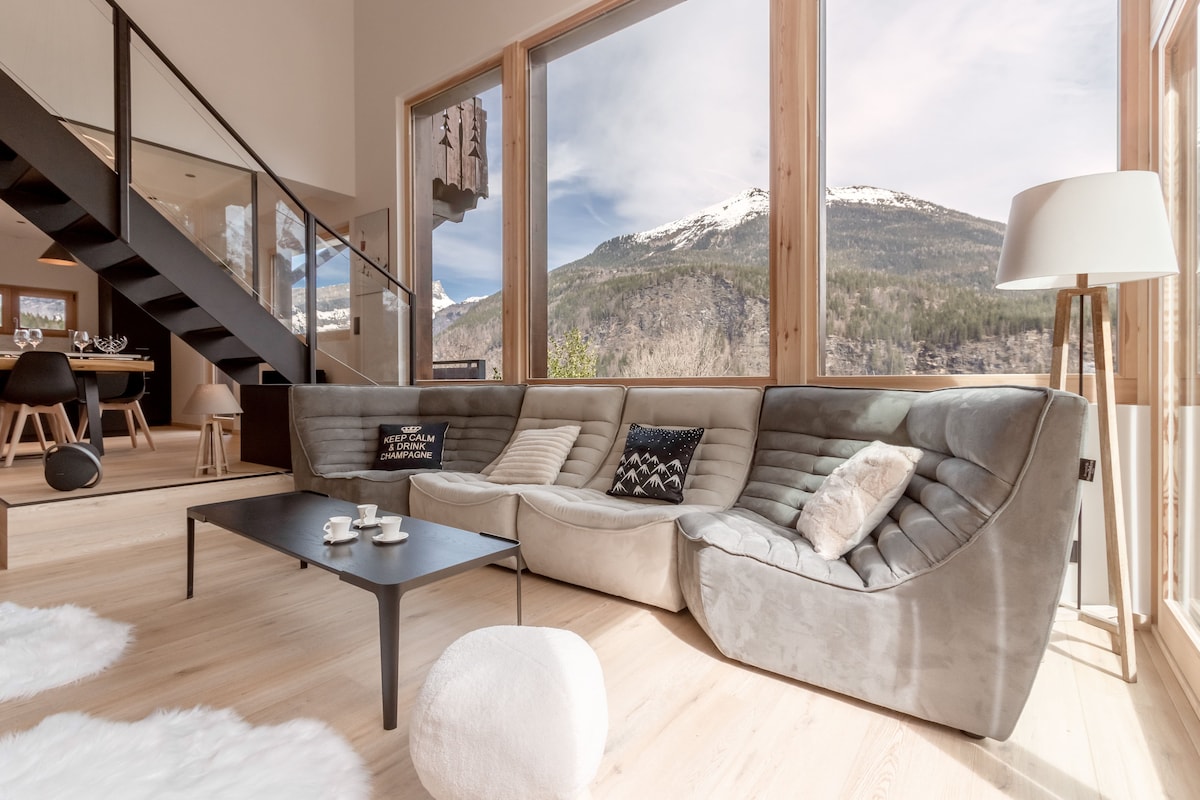 A modern living area features a sectional sofa with a soft grey finish, positioned to face large windows that offer views of the mountains. A low coffee table sits in front, accompanied by two cups and a small decorative pillow. Natural light enhances the airy atmosphere.