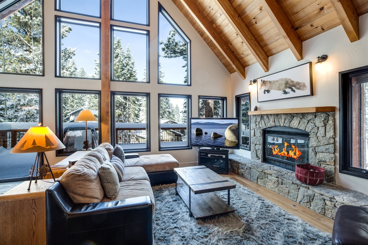 The living room features vaulted ceilings and large windows that enhance natural light and views of the snowy landscape. A comfortable sectional sofa and a rustic coffee table are centered around a stone fireplace, creating a cozy gathering space.