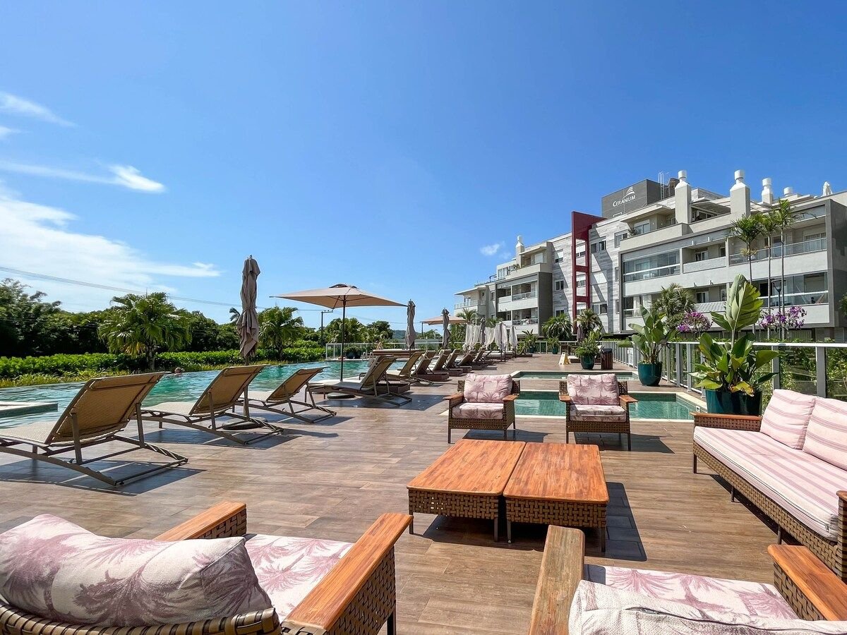 An outdoor relaxation area features comfortable lounge chairs arranged around a serene pool. Square tables made of wood provide space for seating, surrounded by lush greenery and tall palm trees. The modern building in the background houses multiple balconies, contributing to a tranquil atmosphere.