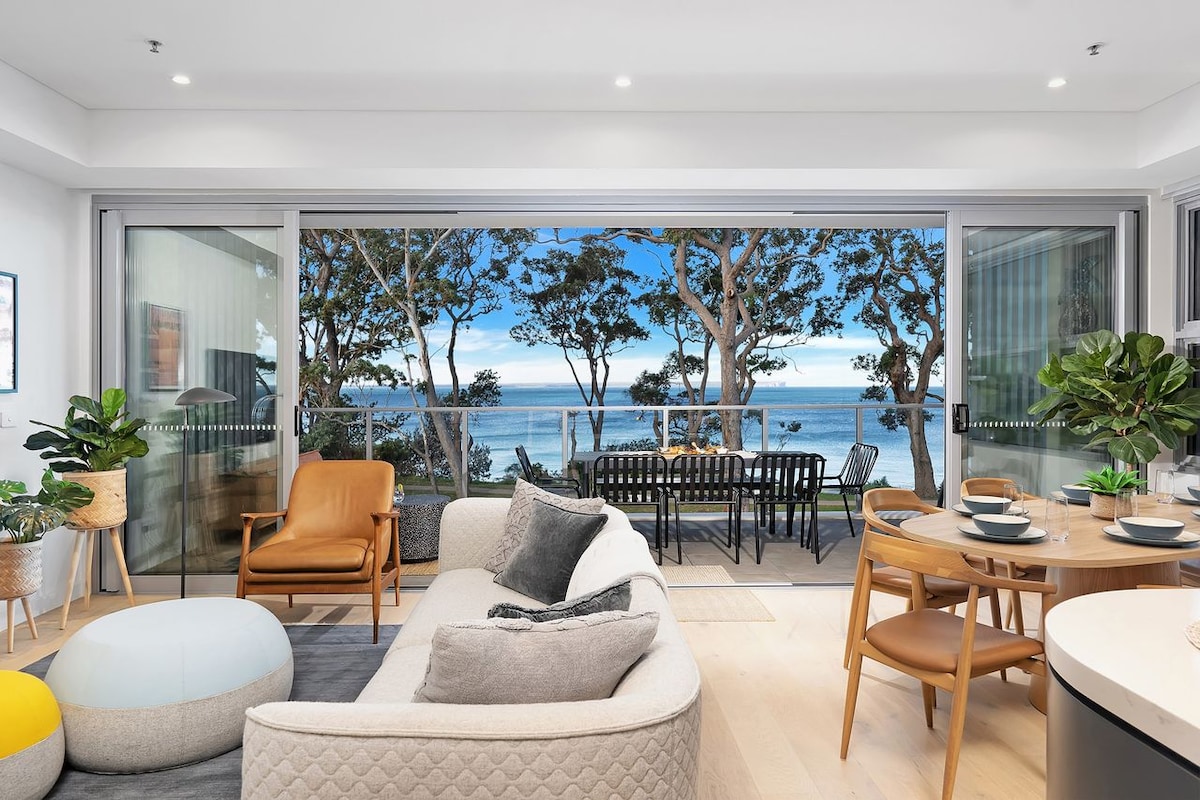 An open-concept living and dining area features a spacious lounge with a light-colored sofa and an armchair. A dining table for six is positioned beside large glass doors, offering a view of the ocean and trees beyond. Natural light brightens the space.