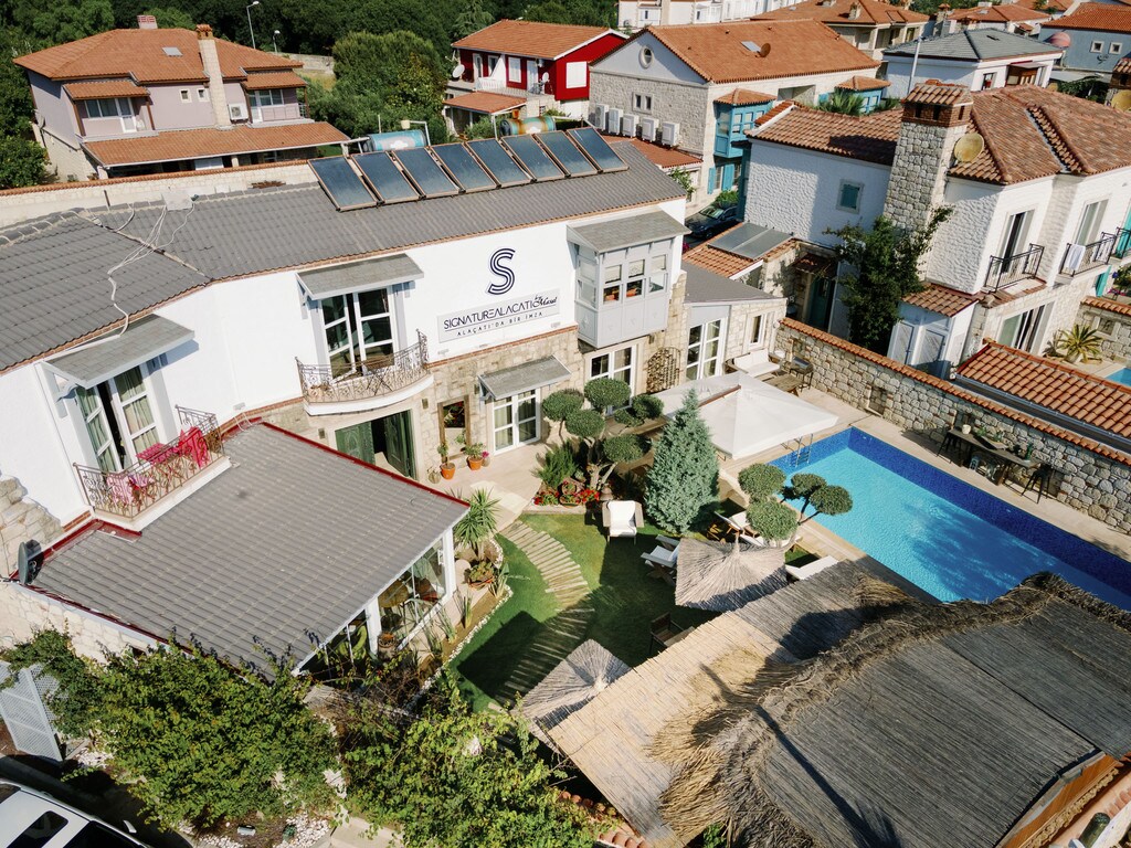 Aerial view of a hotel featuring modern architecture with a large pool and landscaped garden. The property includes seating areas surrounded by greenery and sun loungers by the pool. Nearby, traditional stone houses are partially visible, supporting a charming community atmosphere.