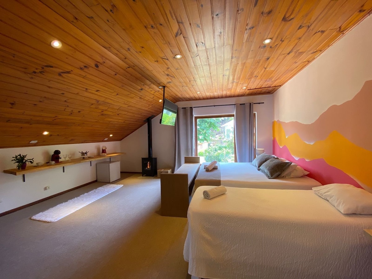 A spacious attic room features a wooden ceiling and natural light flowing through large windows. Two beds are situated beside a modern TV, while a cozy fireplace adds warmth. A minimalist wooden desk is positioned against the wall, enhancing the room's functionality.
