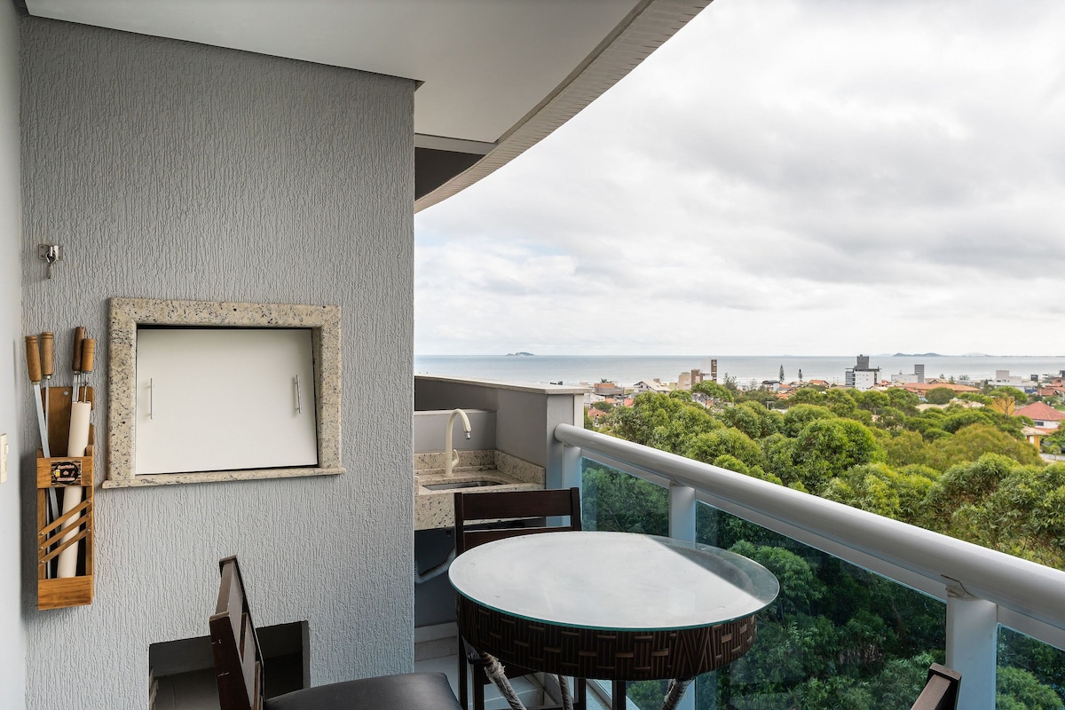 A private balcony features an outdoor dining table and two chairs, with a built-in barbecue area along the wall. Scenic views of the surrounding greenery and ocean are visible beyond the railing, creating a serene outdoor space.