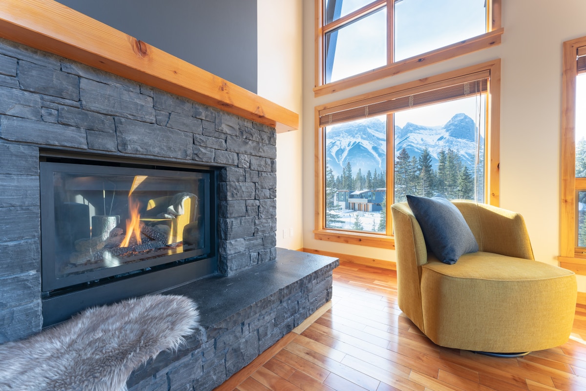 A modern living space features a sleek stone fireplace and a cozy yellow armchair that invites relaxation. Large windows provide panoramic views of the surrounding mountains, while warm wood flooring complements the natural light streaming in.
