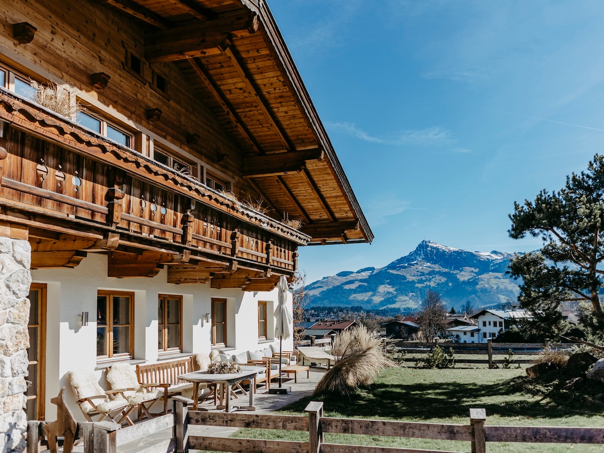 The exterior of the chalet displays a combination of wooden architecture and stone detailing, featuring a spacious terrace with wooden seating. The surrounding landscape includes green fields and distant mountains, all under a clear blue sky.