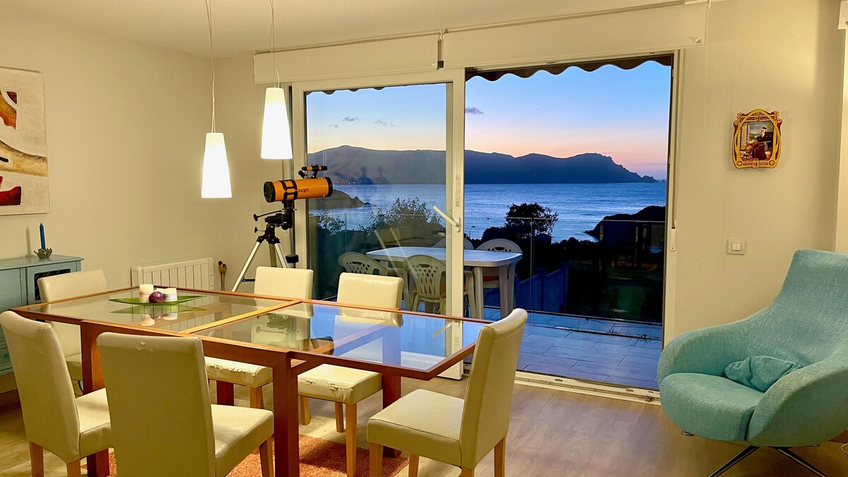 An inviting dining area features a wooden table surrounded by six chairs, with large glass doors revealing views of the coastline at sunset. A telescope is positioned nearby, and a cozy armchair adds a pop of color to the space, enhancing its functionality.