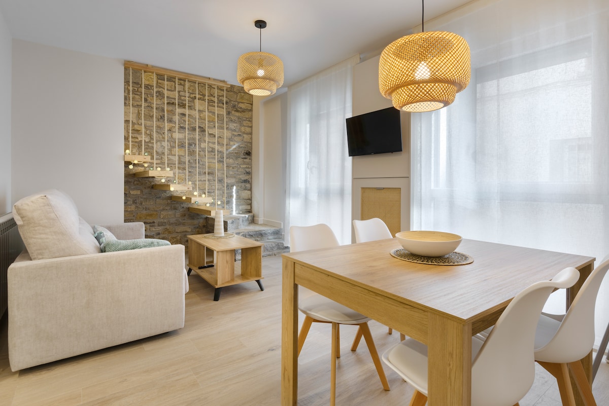 A modern living area features a light-colored sofa and a wooden dining table surrounded by white chairs. Natural light flows through sheer curtains, while the wall is adorned with textured stone. A flat-screen TV is mounted for convenience, and stylish pendant lights provide soft illumination.