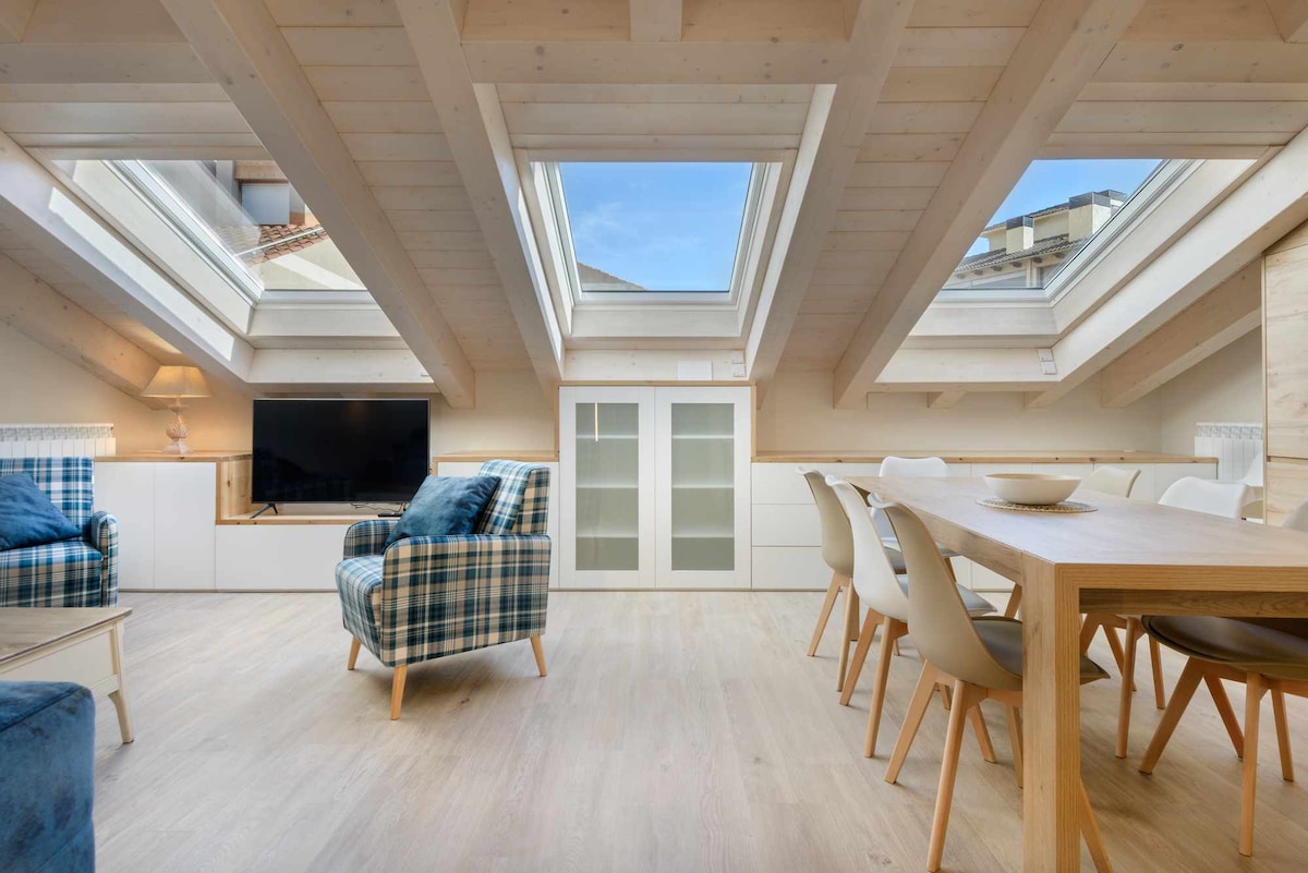 The living area features a cozy seating arrangement with two patterned chairs, complemented by a wooden dining table surrounded by white chairs. Large skylights allow natural light to fill the space, highlighting the wooden beams of the ceiling.