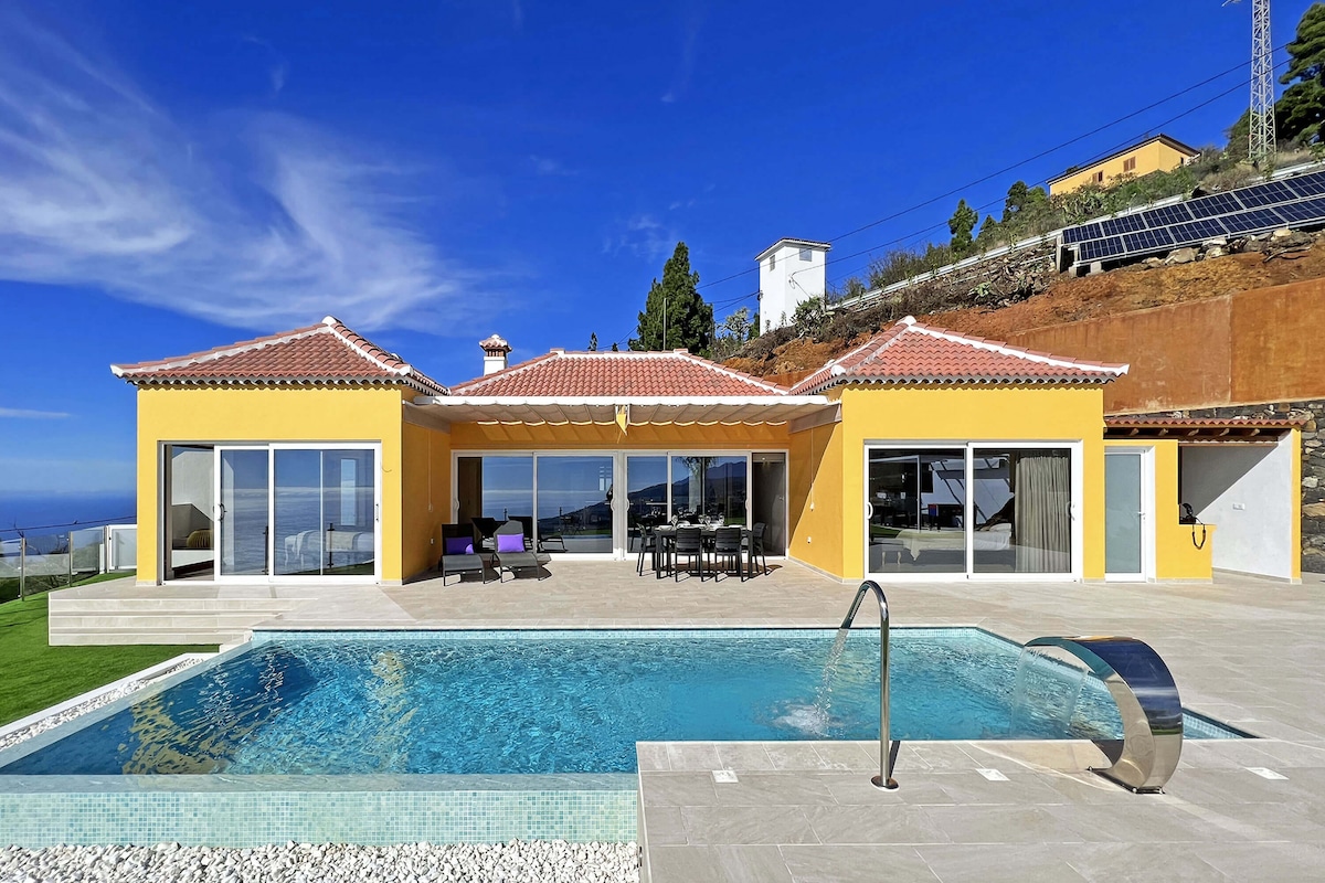 A modern villa with a vibrant yellow exterior is highlighted by large glass doors opening to a spacious patio. A private swimming pool reflects the clear sky, surrounded by clean landscaping. The outdoor dining area is visible, offering a picturesque view of the surrounding area.