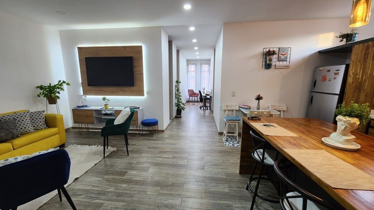 A spacious living area features a yellow sofa and a wooden dining table, accommodating six chairs. A large television is mounted on the wall. Natural light fills the space through large windows, enhancing the inviting atmosphere. Greenery decorates the room, adding freshness.