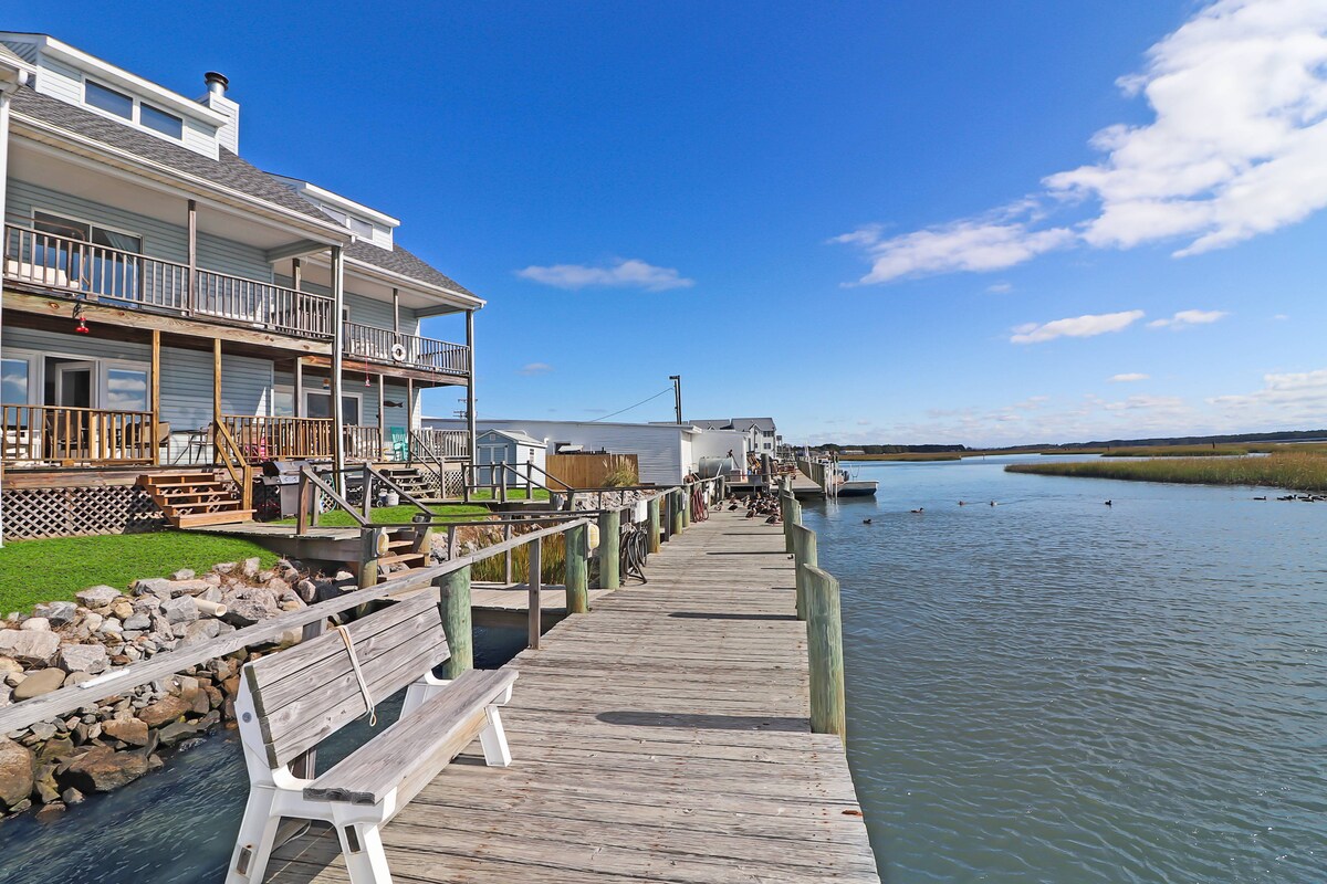A wooden dock extends along the water, bordered by the townhome on one side and marshland beyond. Benches offer seating to enjoy the views. The sky is clear with fluffy clouds, and the calm water reflects the light, creating a serene atmosphere.