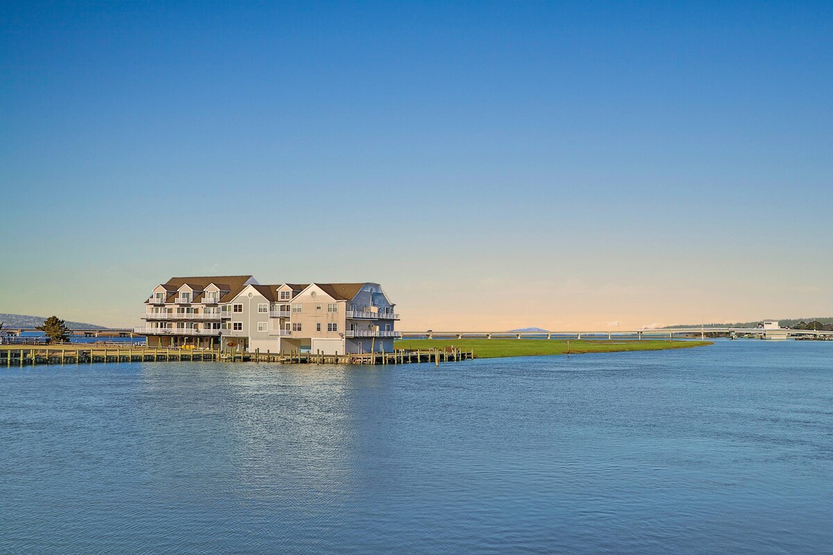A waterfront view captures a multi-level townhome complex situated along the edge of Chincoteague Bay. The calm water reflects the soft hues of the sky, while lush green grass frames the property, creating an inviting scene of tranquility.
