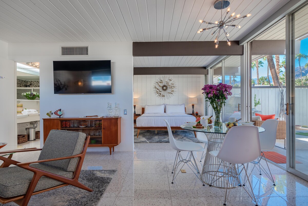 A modern living area features a spacious bed with patterned bedding and a mid-century style side table. A smart TV is mounted on the wall, while a dining area showcases a circular table with white chairs. Natural light enters through large glass doors, leading to an outdoor space.