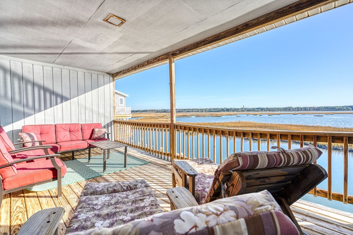A spacious waterfront deck is shown, featuring comfortable seating arrangements including a red sofa and lounge chairs with floral patterns. The serene water view is visible, along with a clear sky, enhancing the calming atmosphere of the outdoor space.