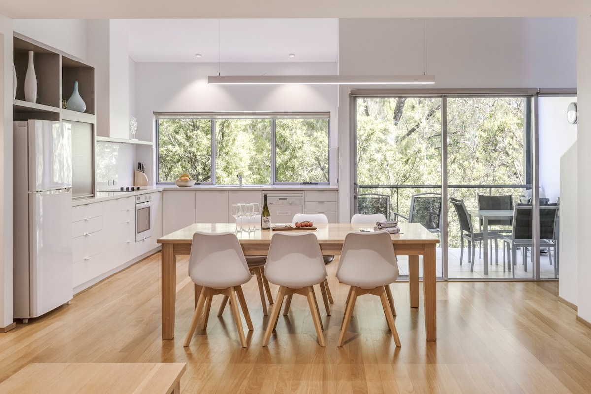 An open-plan kitchen and dining area feature a wooden table surrounded by white chairs. Natural light floods through large windows, highlighting the modern kitchen with stainless steel appliances and cabinetry. A balcony with outdoor seating is visible through the glass doors, creating a seamless connection to nature.