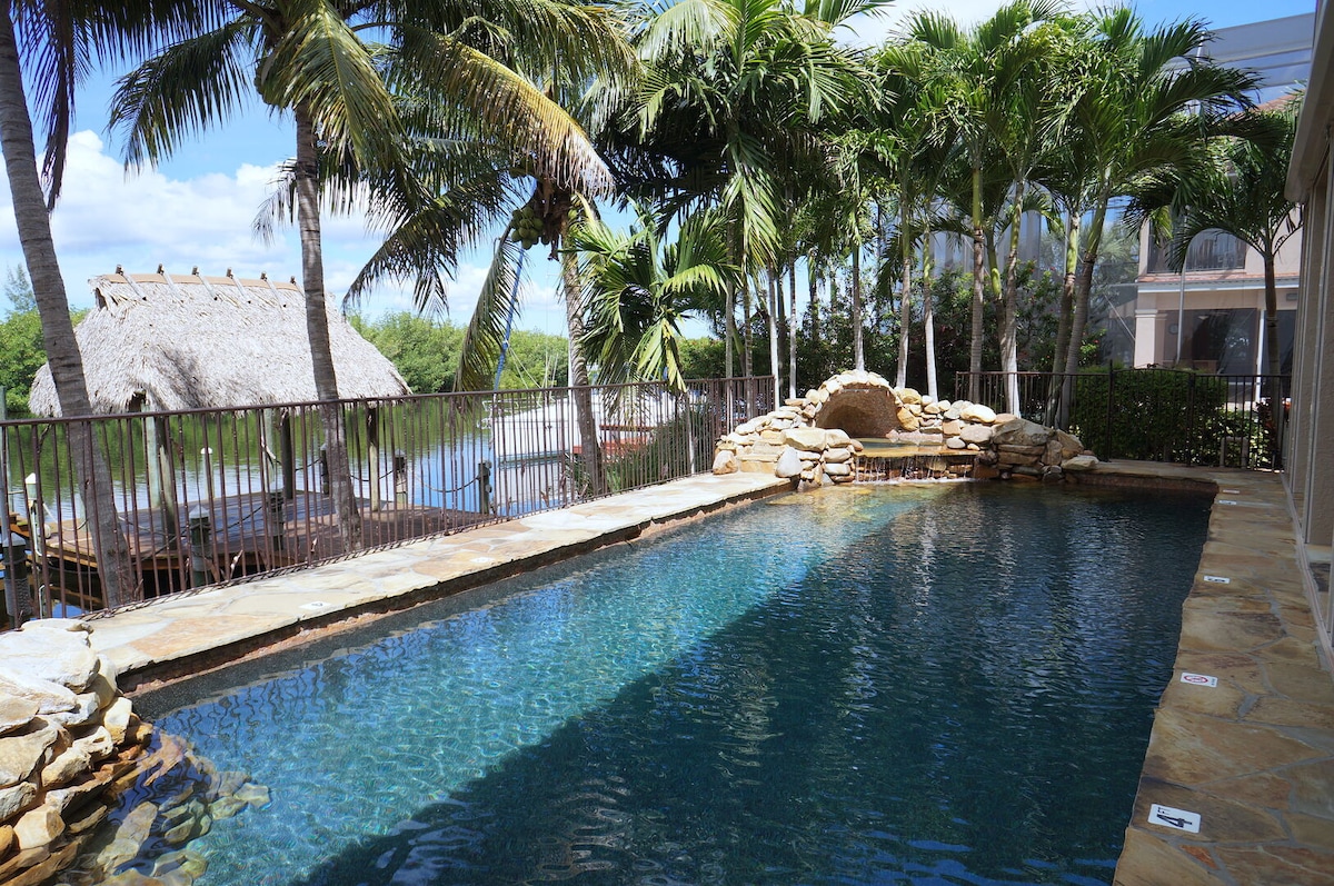 A tranquil pool area is surrounded by lush palm trees, creating a serene environment. Natural stone accents frame the pool, while a thatched gazebo is visible in the background, offering an oasis-like feel. Clear water reflects the sky, inviting relaxation.