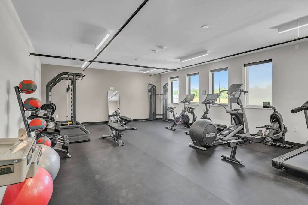 A modern fitness center is equipped with various exercise machines including treadmills and elliptical trainers. Colorful exercise balls rest against the wall, and large windows allow natural light to fill the space. A mirror reflects the room, enhancing its spacious feel.