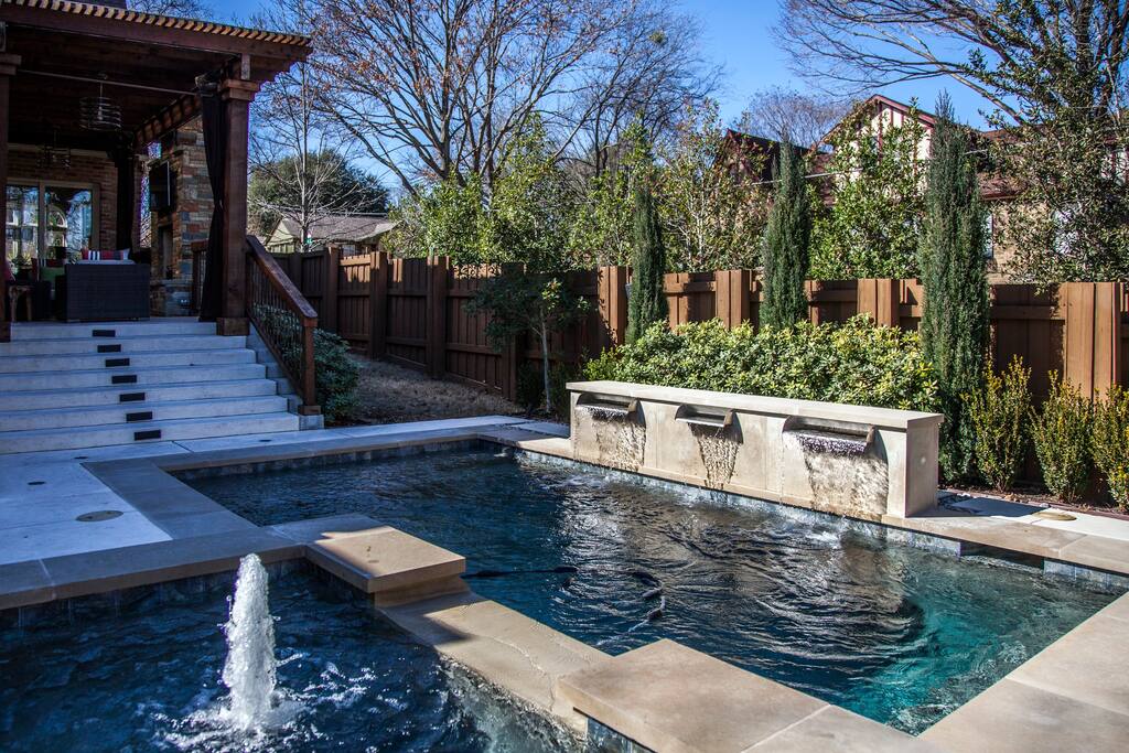 A well-designed outdoor space features a saltwater swimming pool with gentle fountains adding movement to the water. A tranquil spa area is integrated beside the pool, and lush greenery surrounds the perimeter, complemented by carefully maintained trees and shrubs.