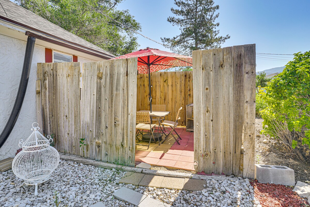 A fenced outdoor patio area is presented, featuring a round table set with two woven chairs. An umbrella provides shade above the table. Graveled space surrounds the patio, complemented by shrubs and trees in the background.