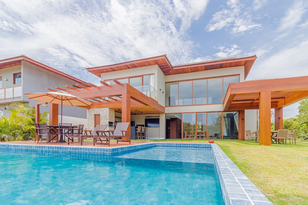 A modern house is depicted with a spacious wooden deck adjacent to the swimming pool, featuring lounge chairs and a shaded area. Large glass windows provide a view of the interior. Lush greenery surrounds the property, contributing to a serene outdoor setting.