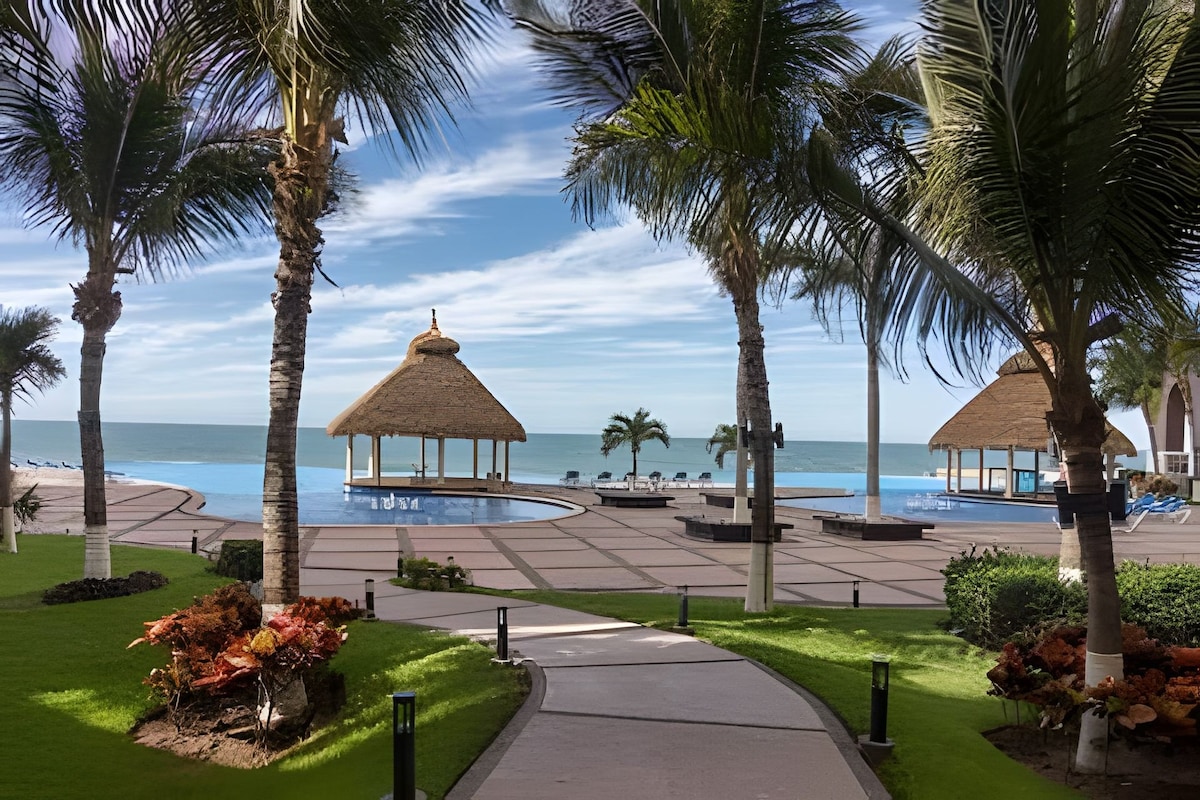 A beautifully landscaped outdoor area features palm trees surrounding a large pool. Thatched-roof cabanas provide shaded seating options. The walkway leads directly to the pool, with the ocean visible in the background under a clear sky.