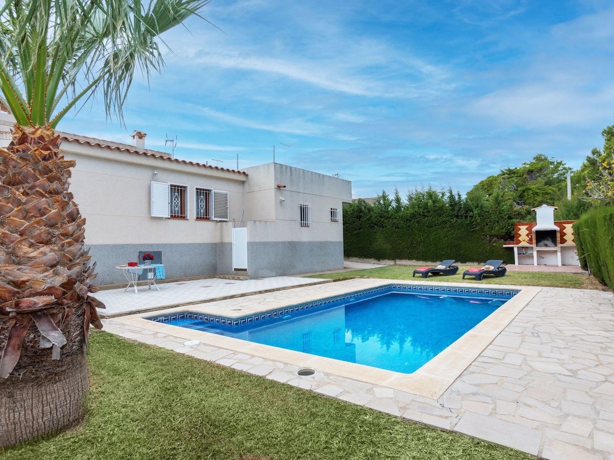 A private swimming pool is set within a well-kept garden, surrounded by lush greenery. A stone patio borders the pool, with lounge chairs positioned for relaxation. A barbecue area is visible beside the villa, providing options for outdoor dining.
