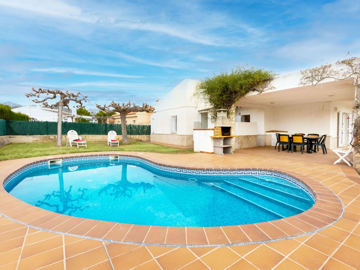 A kidney-shaped swimming pool is surrounded by a tiled patio, featuring lounge chairs and a barbecue area. A covered terrace with dining furniture is visible, offering shaded seating options. The garden features well-maintained vegetation, with trees providing a natural backdrop.