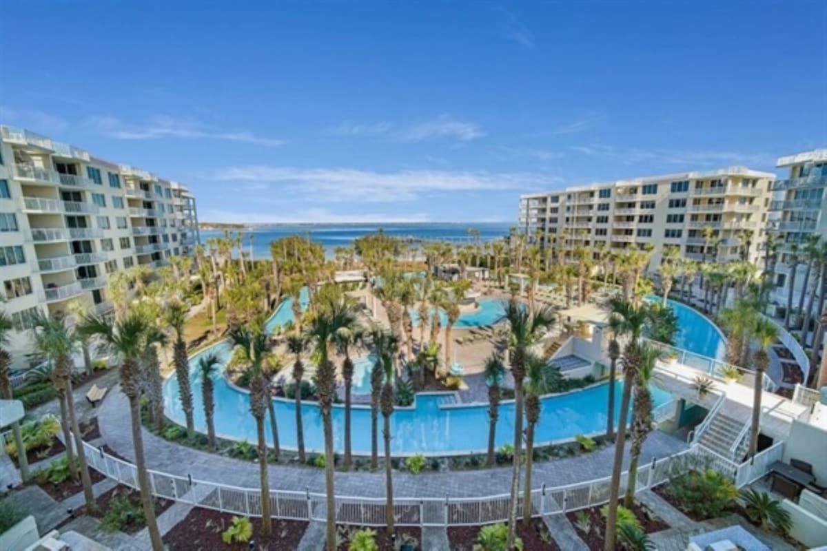 A sweeping view showcases the expansive lazy river pool surrounded by lush palm trees and manicured landscaping. The serene blue of the water contrasts with the bright sky, and buildings are positioned elegantly around the pool area, offering a vibrant resort atmosphere.