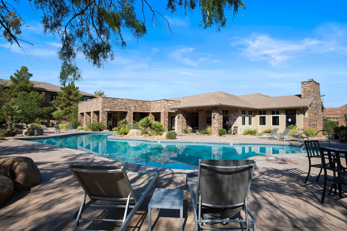 An outdoor pool area is featured with lounge chairs positioned nearby. The stone building provides a backdrop, surrounded by landscaped gardens and desert vegetation. A clear blue sky enhances the inviting atmosphere of the poolside space, ideal for relaxation and enjoyment.