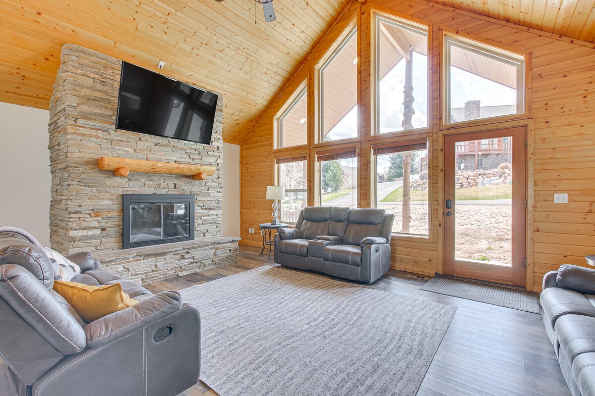 A welcoming living area features a stone fireplace and a large mounted TV. Comfortable seating is provided by two reclining sofas in shades of gray. Floor-to-ceiling windows allow natural light to fill the space, revealing views of the outdoor surroundings.