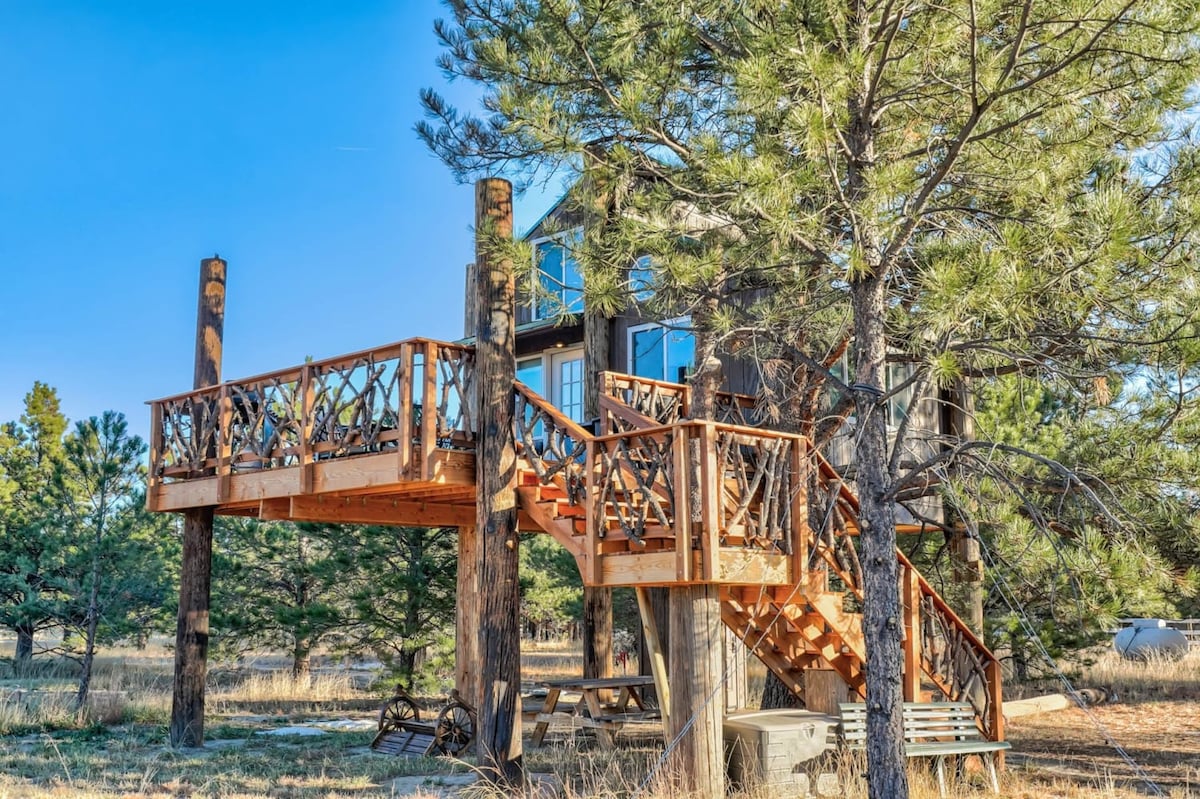 A wooden staircase leads to a spacious deck adorned with rustic railings. The treehouse, nestled among tall pines, features large windows reflecting the natural surroundings and a wooden structure that harmonizes with its environment.