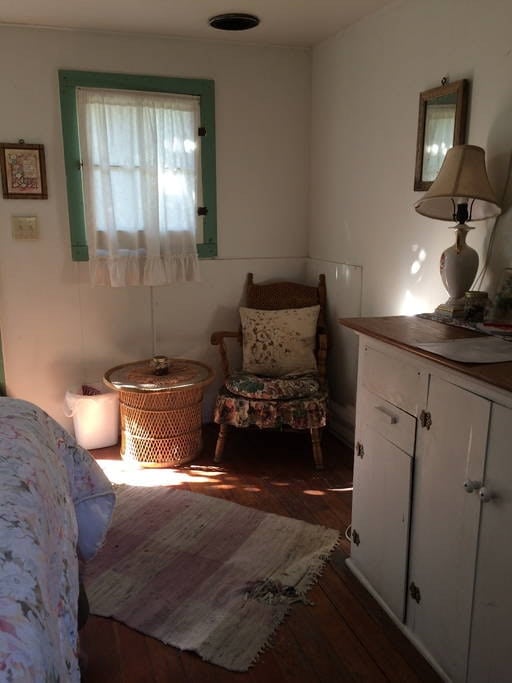 A cozy corner of the cabin features a woven chair with floral upholstery beside a small table topped with a lamp. Natural light filters through a window with sheer curtains, illuminating the warm wooden floor and light-colored walls.