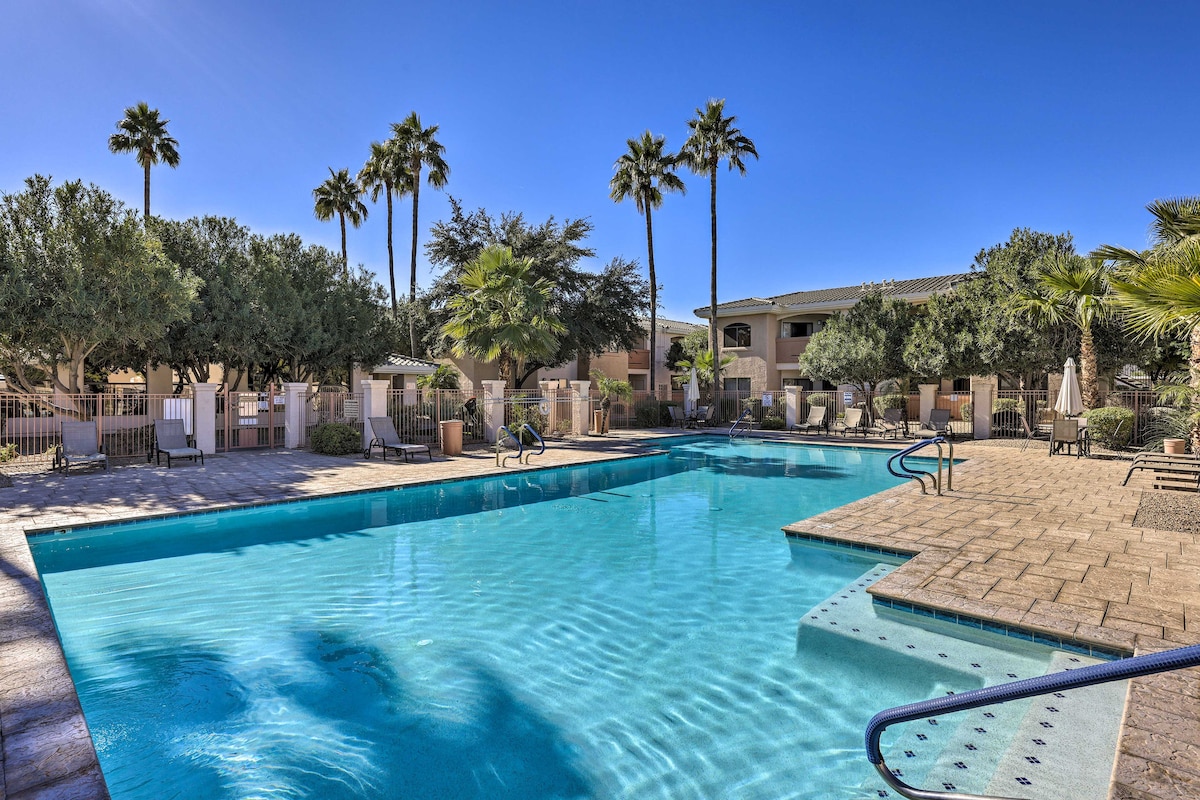 A community pool features clear blue water surrounded by a spacious patio area. Palm trees and shrubs provide greenery, while lounge chairs are arranged along the sides. The pool is framed by a safety fence and is part of a well-maintained complex.