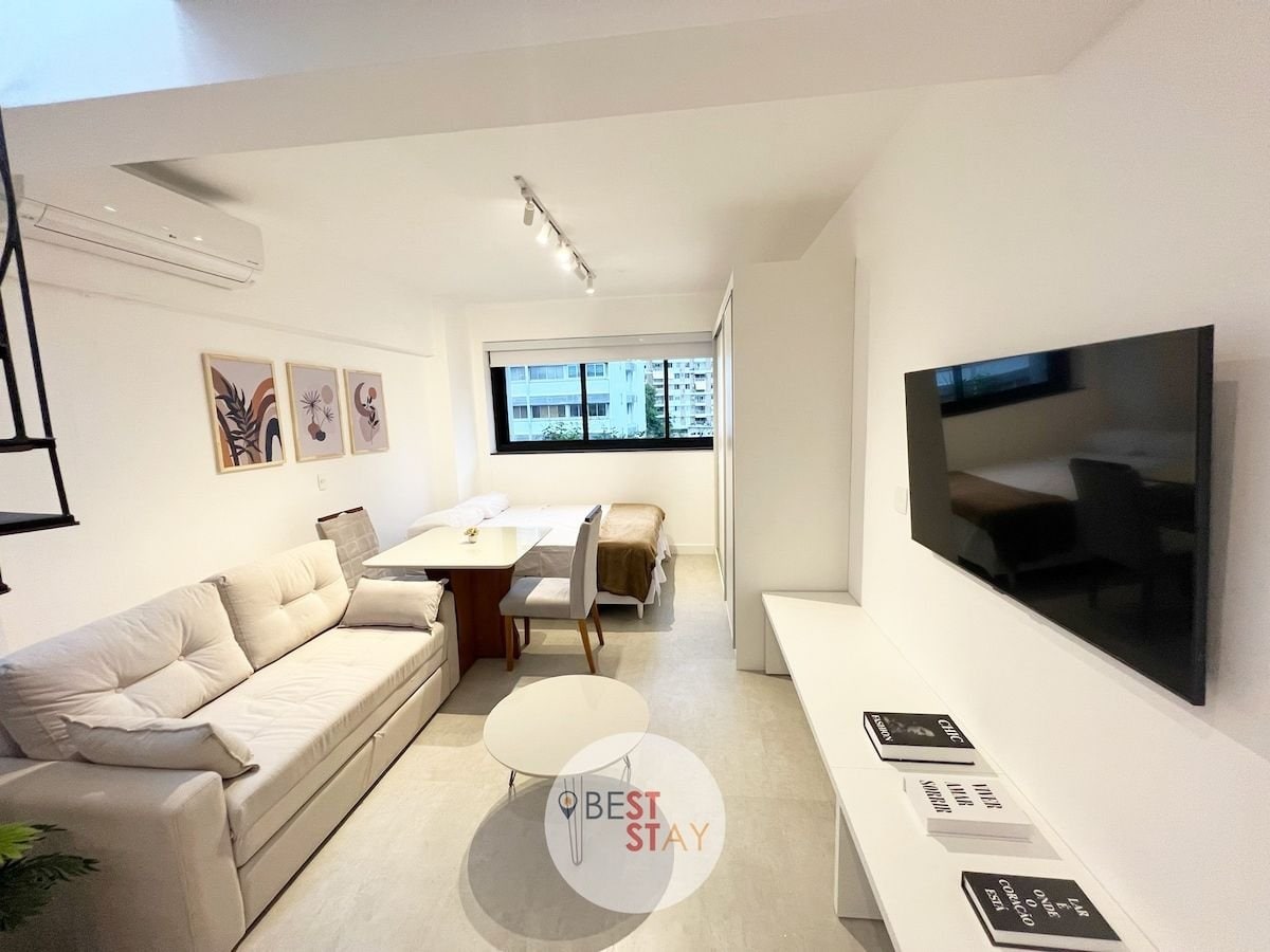 A renovated living space features a light-colored sofa and a coffee table. A smart TV is mounted on the wall, opposite a double bed. Artwork adorns the walls, and natural light fills the room through large windows, enhancing the bright and airy ambiance.
