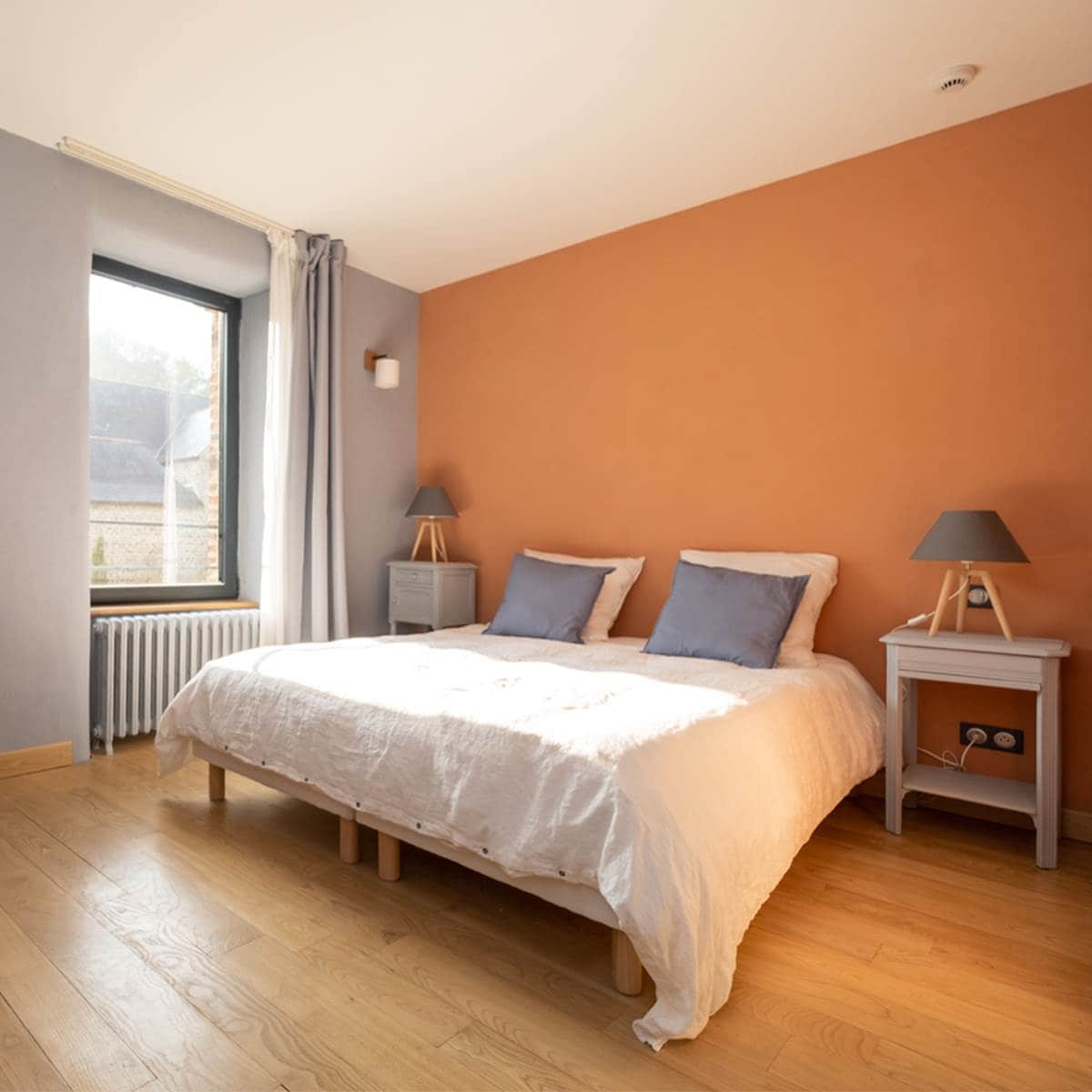 A bedroom features an orange accent wall paired with soft bluish gray walls. A spacious bed is arranged with white linen sheets and two blue cushions. Nightstands with lamps are positioned on either side, and natural light filters in through a large window.