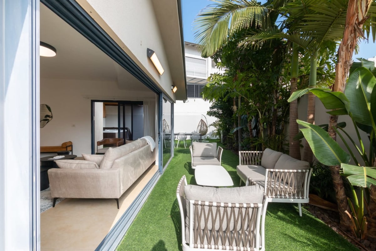 A spacious outdoor area is framed by tall greenery, featuring contemporary seating arrangements on a grassy surface. Light filters through a large opening from the interior, highlighting a modern lounge space within. The atmosphere is serene, inviting relaxation in the natural surroundings.