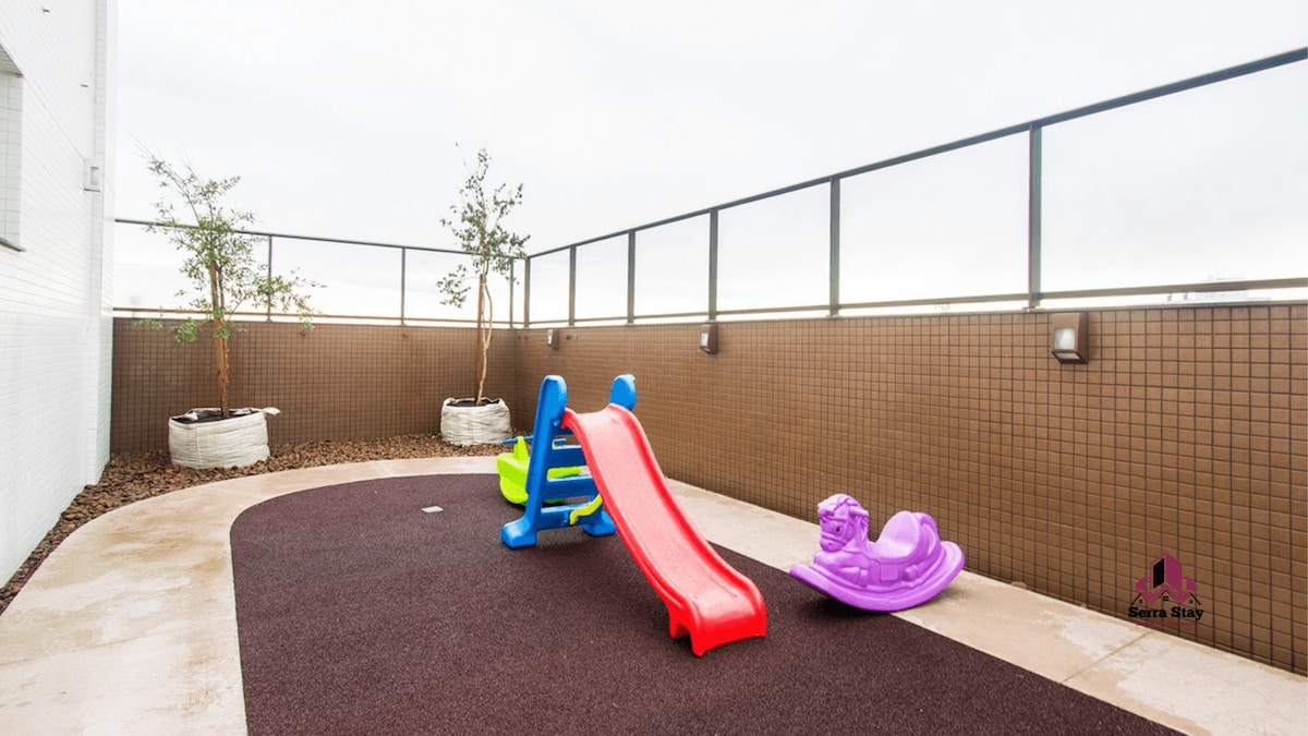 A shared recreation area on the rooftop features a colorful playground with a small slide and a rocking toy. Potted plants are placed along the perimeter, and safety surfacing covers the ground, providing a secure space for children to play.