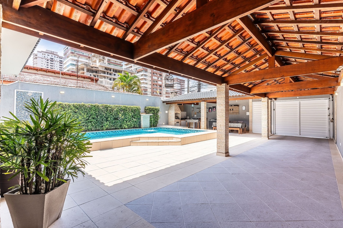 An outdoor space features a spacious patio area with a stunning pool surrounded by greenery. A wooden roof offers shade, while seating arrangements for gatherings are present. The surrounding walls are adorned with plants, providing a tranquil setting for relaxation and enjoyment.