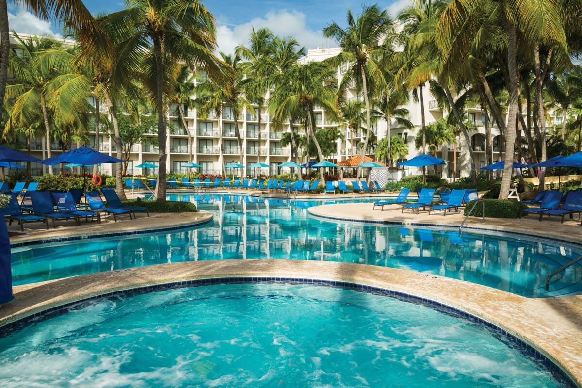 A large outdoor pool is surrounded by lush palm trees and sun loungers with blue umbrellas. The tranquil water reflects the clear sky, creating a serene atmosphere. The pool area is designed for relaxation and enjoyment, featuring distinct zones for swimming and lounging.