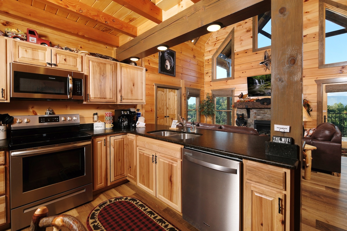 The fully equipped kitchen features stainless steel appliances, including a microwave and dishwasher, against natural wood cabinetry. A dual sink integrates with granite countertops. Ample natural light streams through the large windows, providing a view of the surrounding mountains.