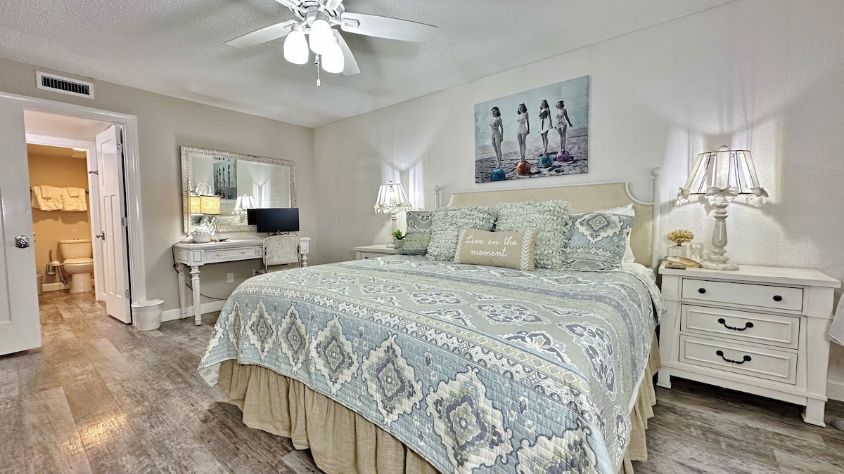 A spacious bedroom features a King bed adorned with a patterned quilt, complemented by matching bedside lamps. A decorative piece with three figures at the beach hangs above the bed. A work desk and a smart TV are situated nearby, with an en-suite bathroom visible through an open door.