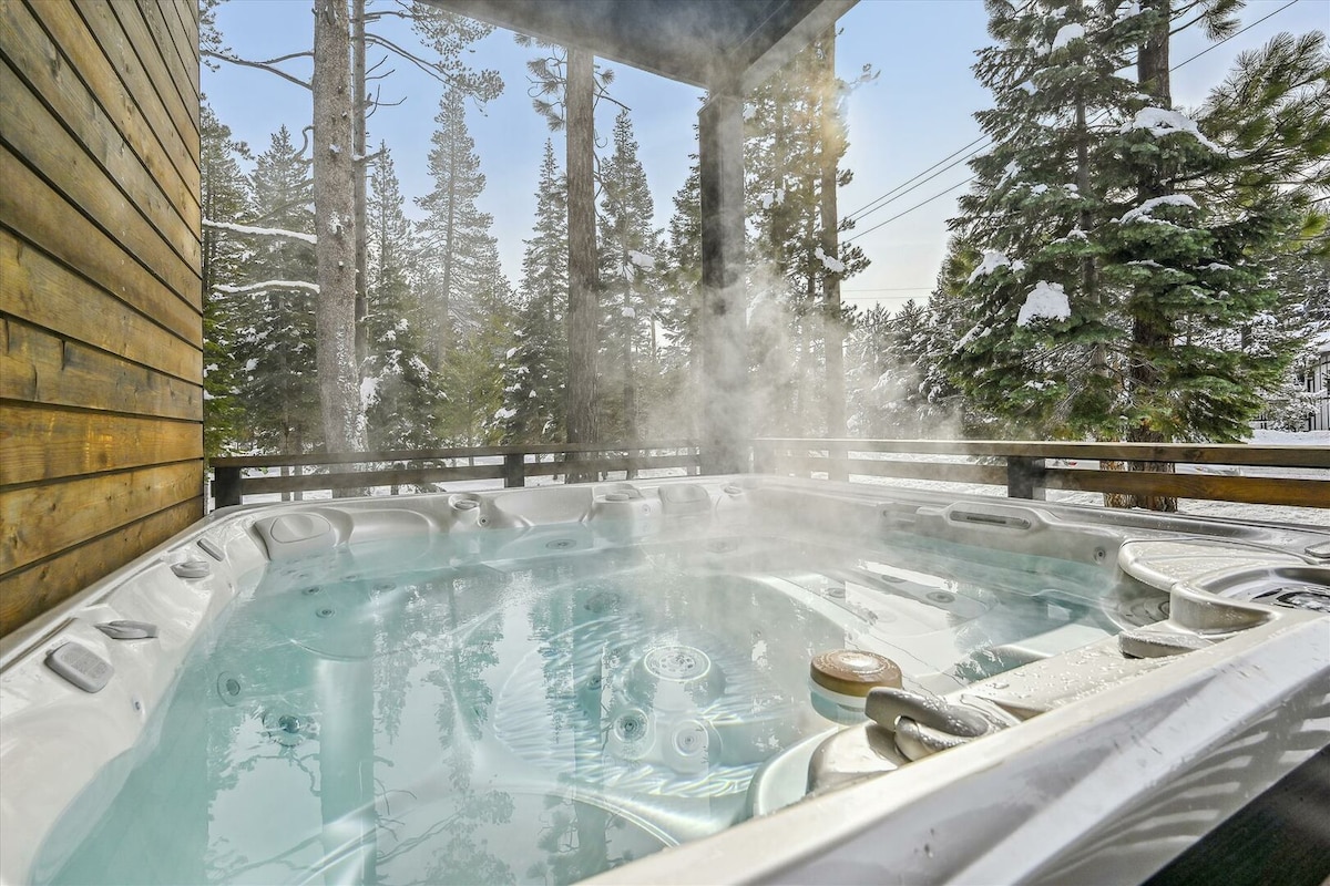 A hot tub is situated on a covered deck, with steam rising into the crisp winter air. Surrounding evergreen trees provide a natural backdrop, and the wooden panels of the deck create a cozy, secluded environment.