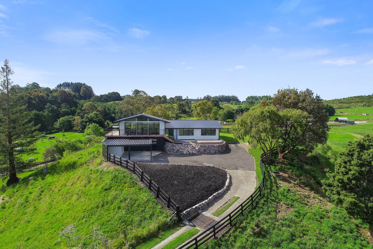 The exterior view showcases a spacious four-bedroom home nestled on a hillside, surrounded by lush greenery. A winding path leads up to the home, with a large gravel area in front and a well-manicured lawn. The landscape features distant trees and open farmland.