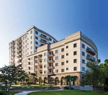 The resort building features a distinct architectural design with multiple floors and numerous balconies. A landscaped area with greenery is visible in the foreground, highlighting the outdoor space. The bright blue sky provides an inviting backdrop to this modern structure.